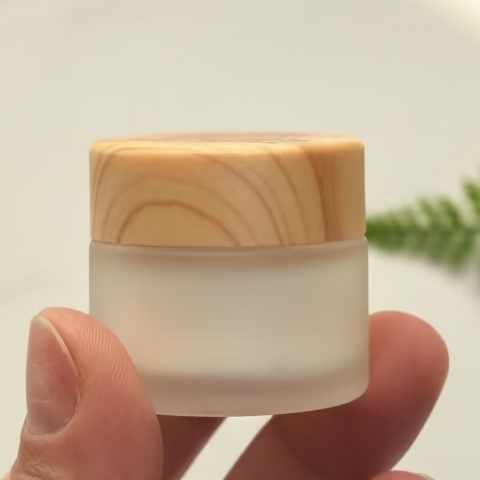 Hand holding a small white jar with a wooden lid against a blurred natural background