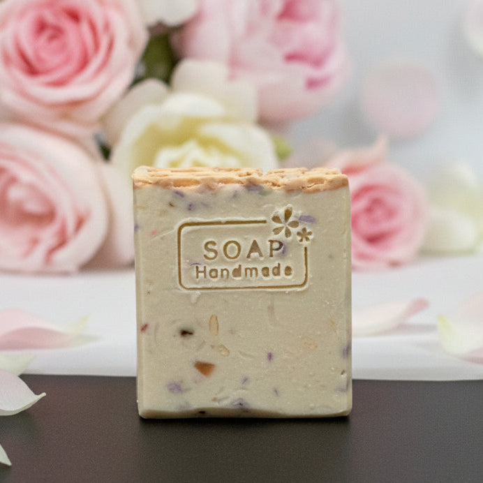 A handmade rectangular bar of soap with a rough, rustic top edge, featuring a stamped label that reads 'SOAP Handmade' with a small flower design. The soap has a light beige base with small brown and purple botanical inclusions.